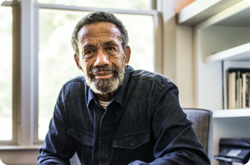 Older gray haired black man sitting by a window in front of a bookshelf